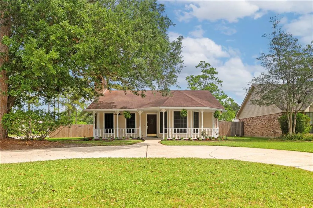 A tan house with a brown roof and white porch sits on a green lawn under a blue sky with trees.