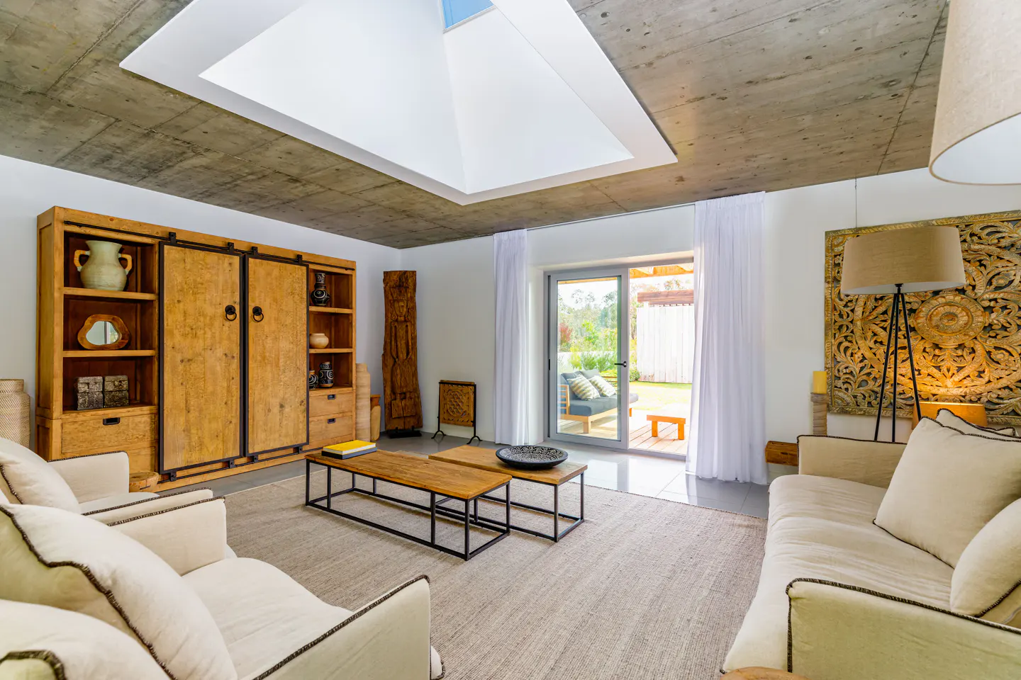 Living room with white sofas, wood cabinets, and a concrete ceiling with a skylight. Sliding glass doors lead to a green lawn.