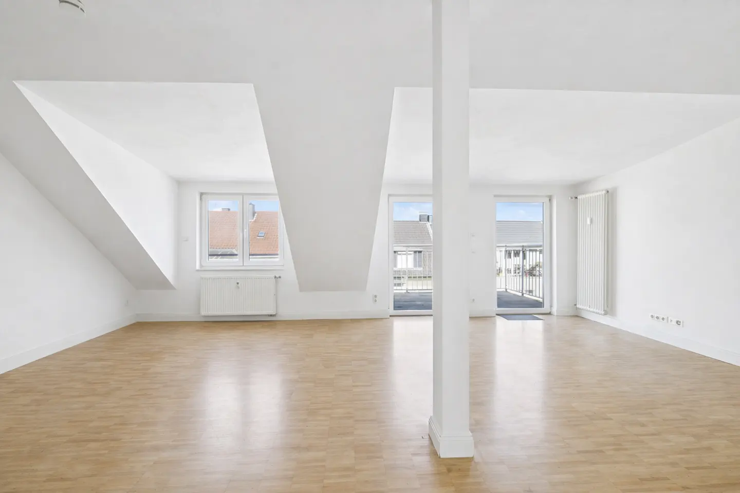 Bright, empty attic apartment with light wood floors, white walls, windows, and a support column.