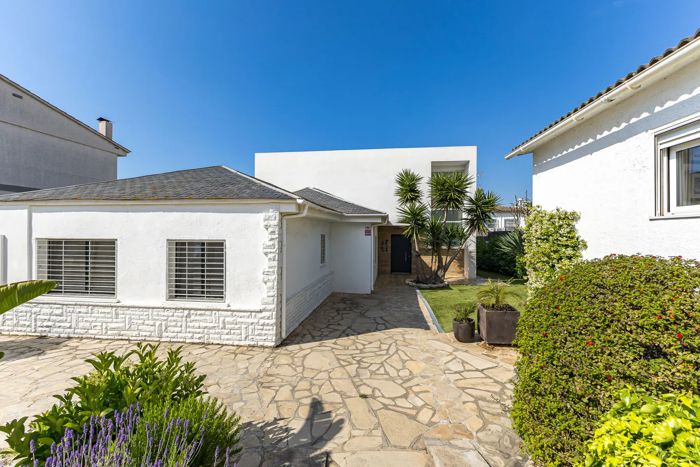 Exterior view of a white, modern house with a stone driveway, green plants, and a clear blue sky.