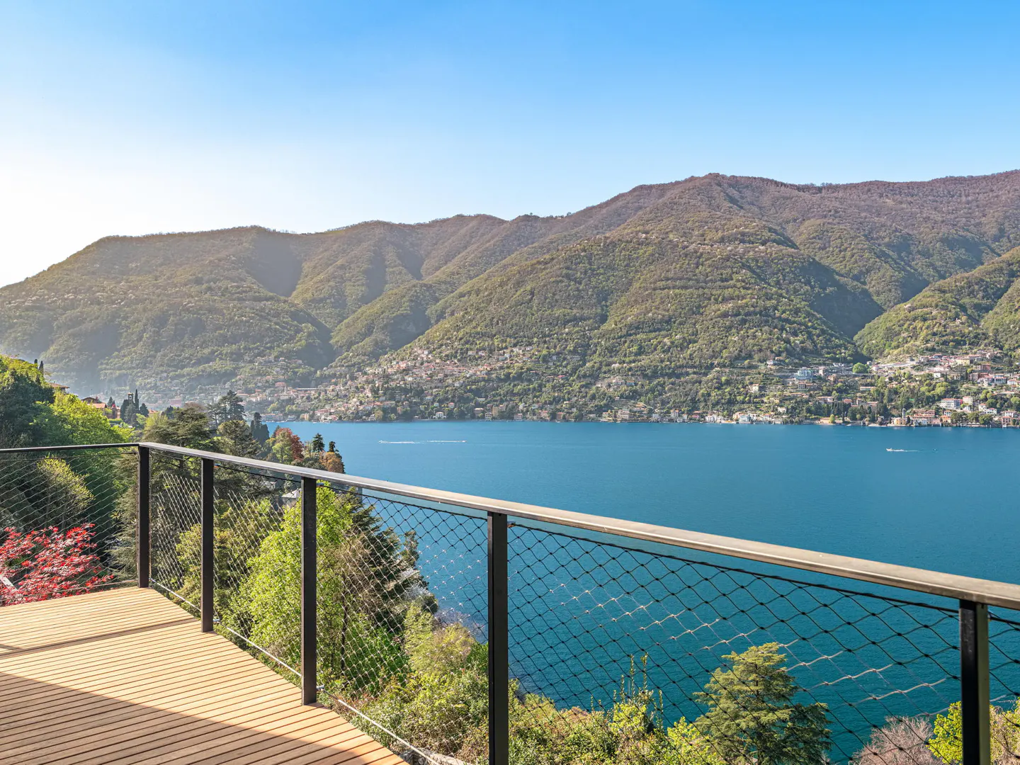 Scenic view from a wooden deck with a metal railing overlooking a blue lake and green mountains under a clear blue sky.