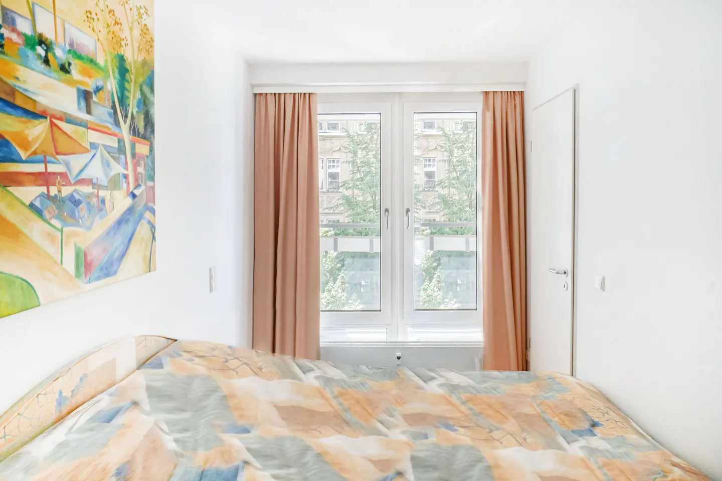 Bedroom with a bed covered in a patterned quilt, a window with peach curtains, and an abstract painting on the wall.