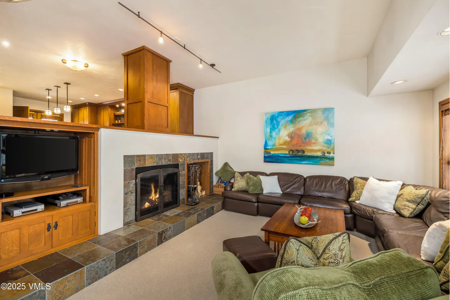 Living room with a brown leather sectional, a stone fireplace with a fire, and a TV in a wooden cabinet. An abstract painting hangs on the wall.