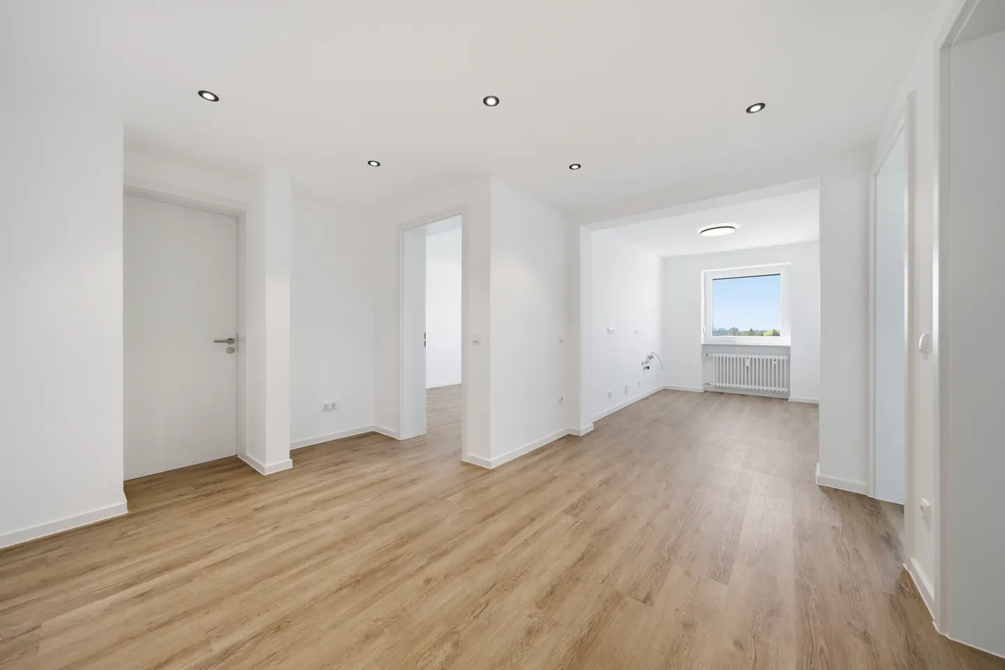Bright, empty room with light wood floors, white walls, and recessed lighting. A window shows a view of trees and sky.
