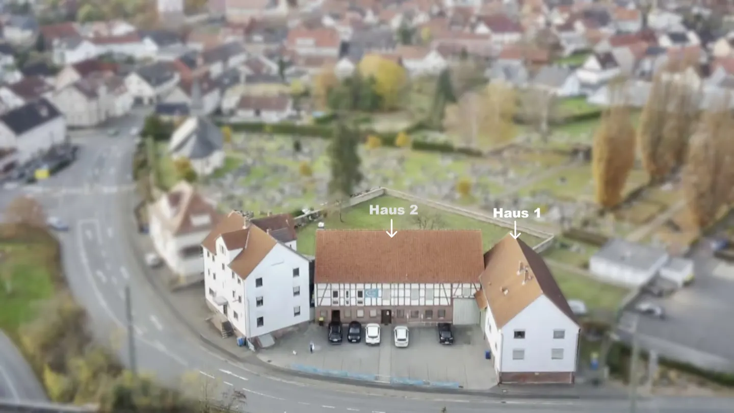 Aerial view of two white buildings with brown roofs, labeled Haus 1 and Haus 2, with cars parked in front. A cemetery is in the background.