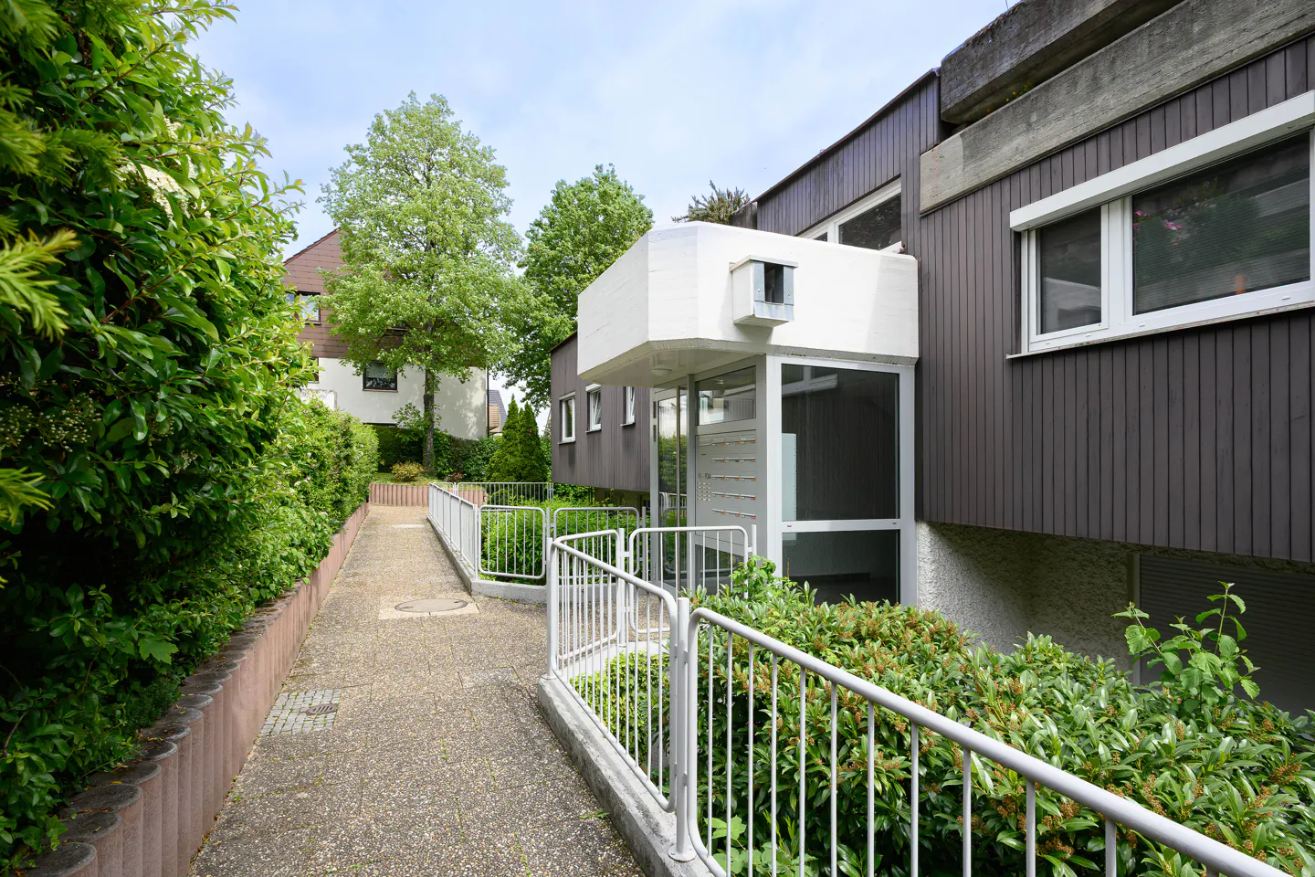 Exterior view of a brown building with white trim, a walkway, and green foliage.
