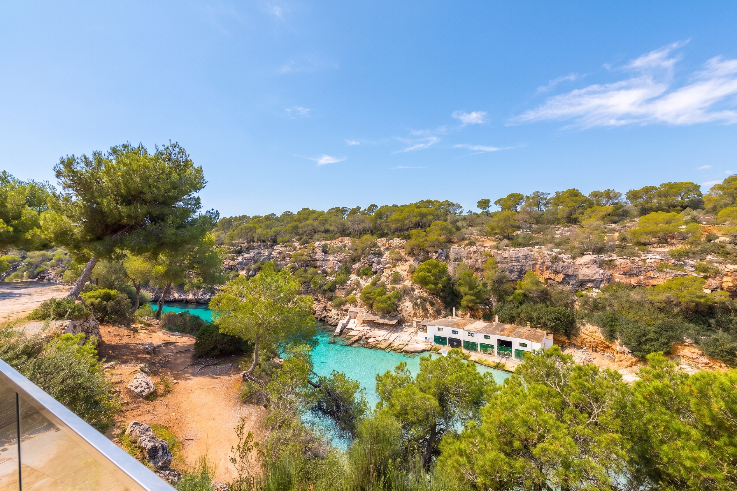 Scenic view of turquoise water inlet surrounded by green trees and rocky cliffs under a blue sky. A white building sits on the water's edge.