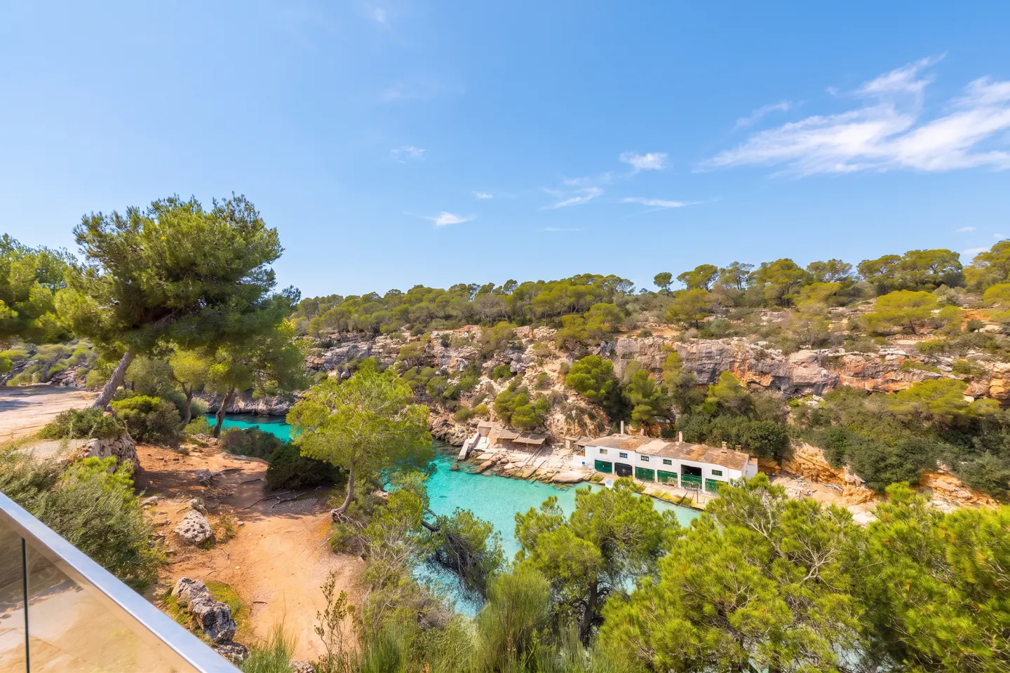 Scenic view of turquoise water inlet surrounded by green trees and rocky cliffs under a blue sky. A white building sits on the water's edge.