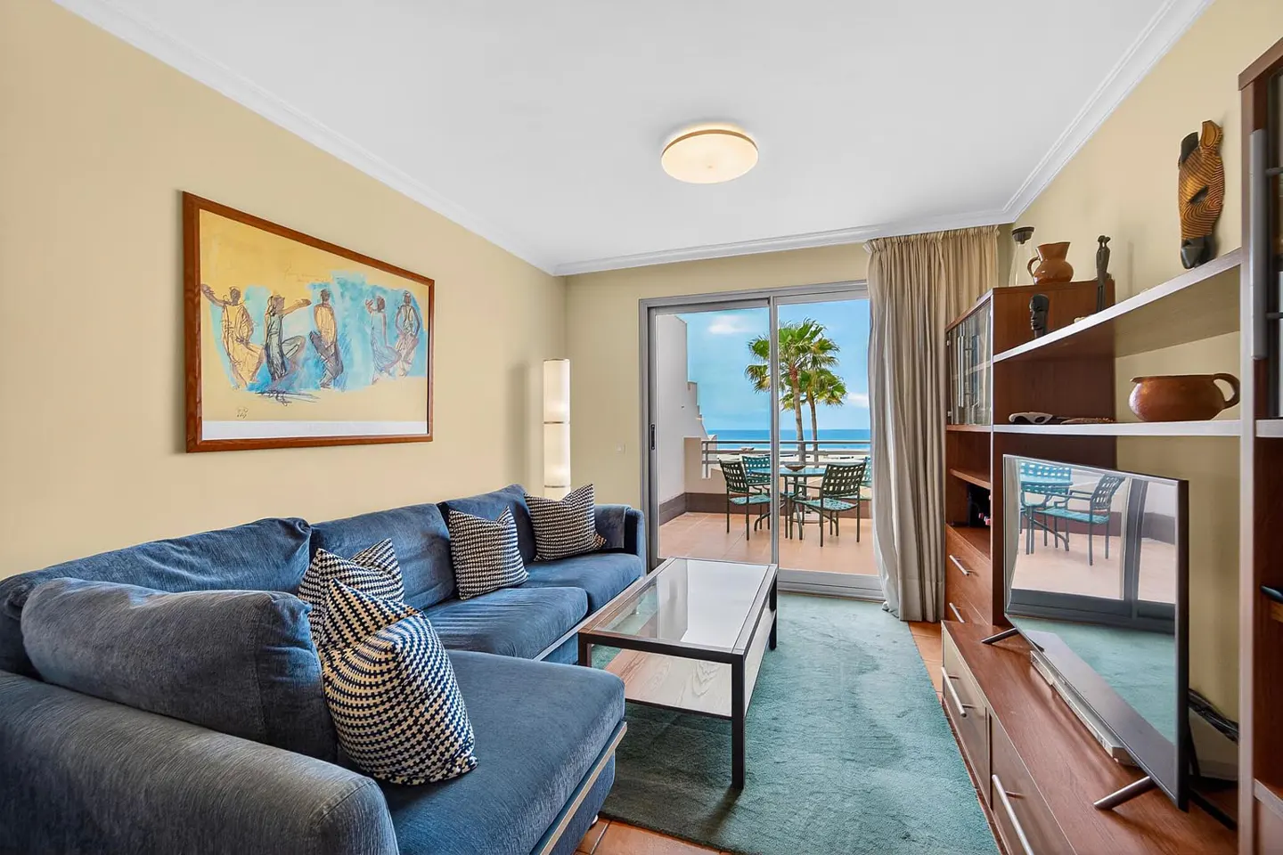 Living room with blue sectional sofa, glass coffee table, and ocean view from the balcony. Artwork on the wall.