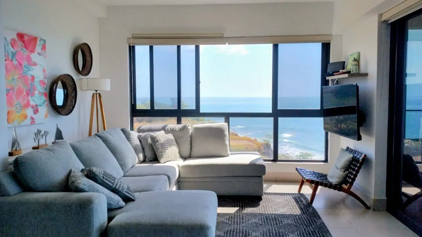 Bright living room with ocean view. Gray sectional sofa, floral art, and large windows overlooking the blue ocean. A TV and chair are on the right.