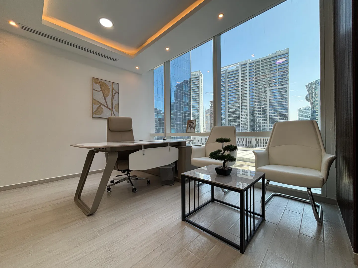 Bright office space with a desk, chair, and seating area. Large windows offer a city view. A small bonsai tree sits on a table.