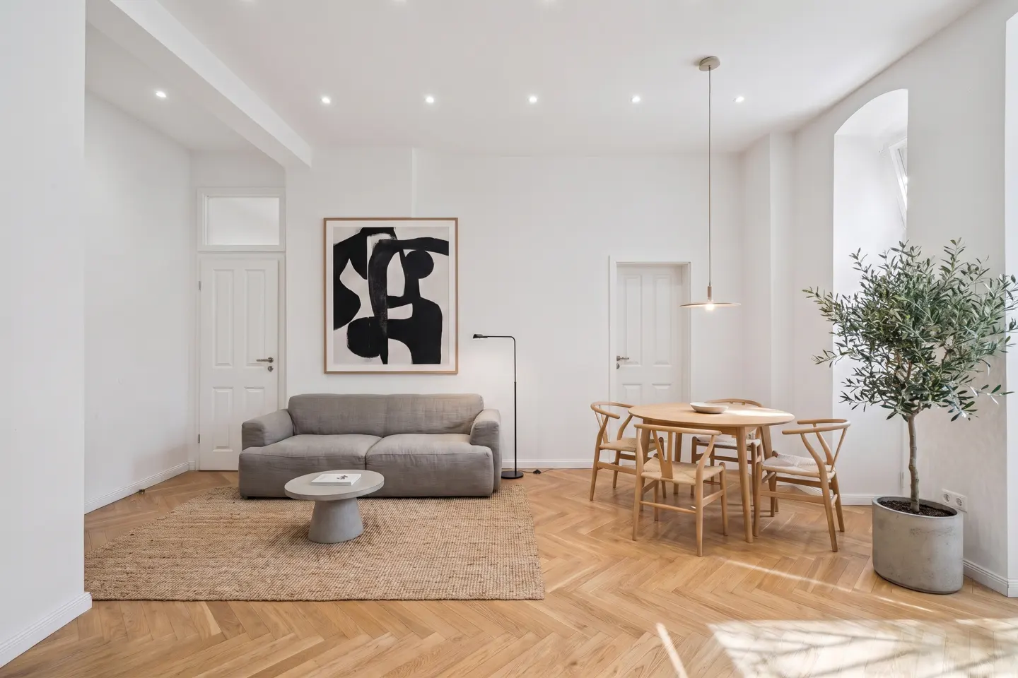 Bright living room with white walls, wood floors, and modern decor. A gray sofa sits under abstract art, near a round table with wooden chairs. A potted olive tree adds a touch of nature.