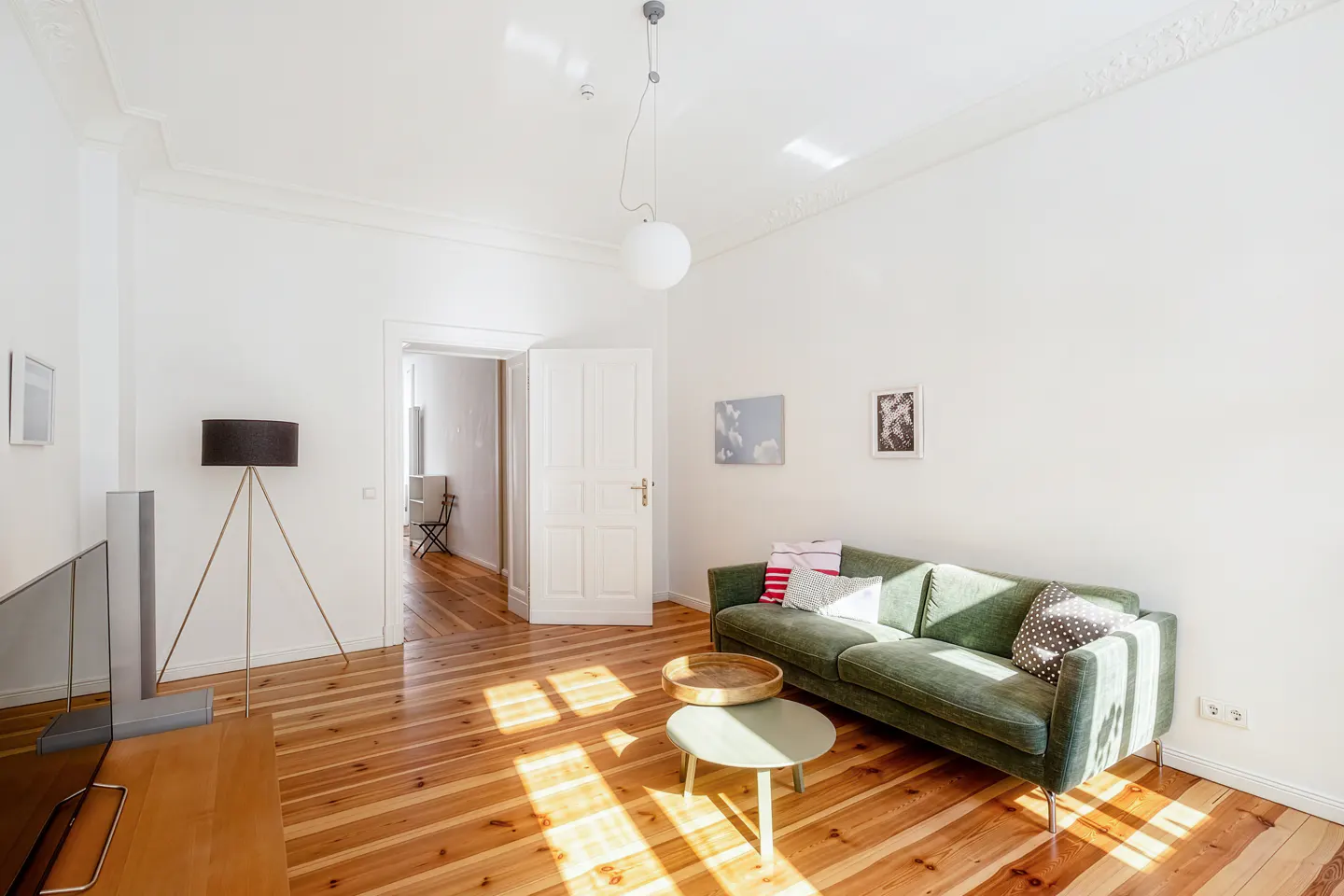 Bright living room with wood floors, white walls, and a green sofa. Sunlight streams in, highlighting a round coffee table and an open doorway.