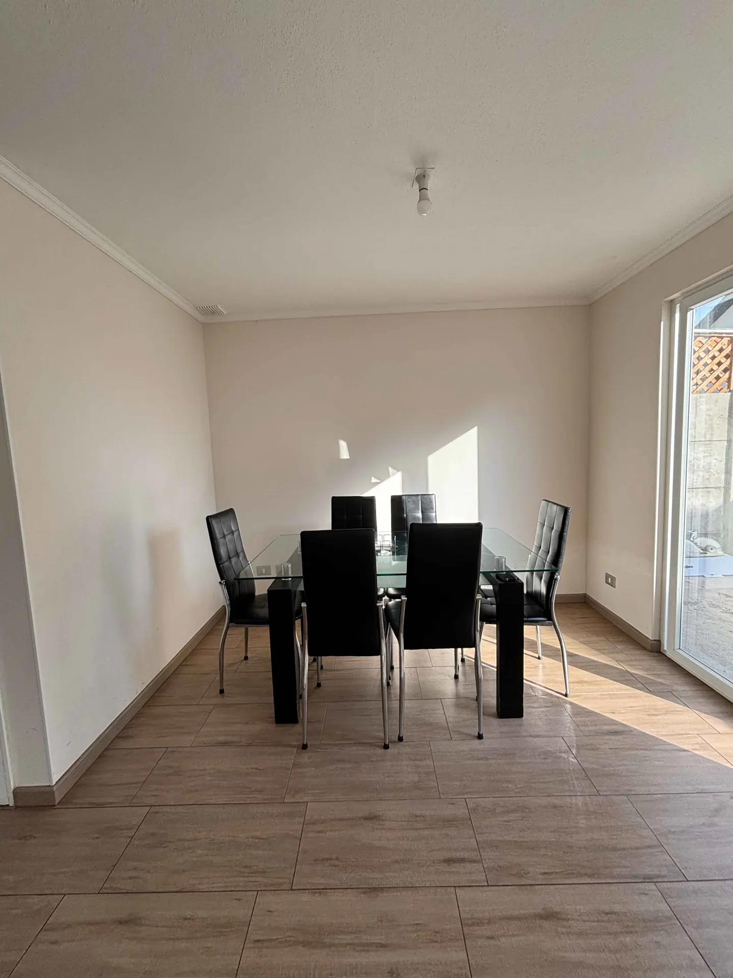A dining room with a glass table and six black chairs on a wood-look tile floor. Sunlight streams in from a sliding glass door.