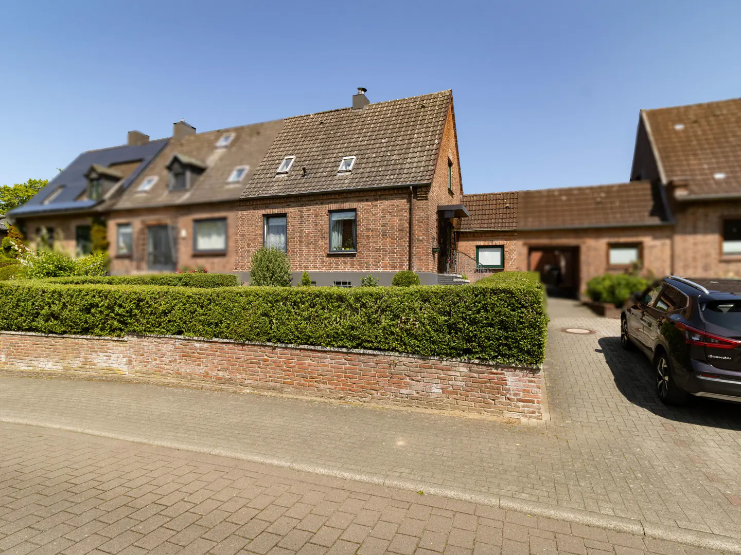 Exterior view of a red brick house with a green hedge, a brick wall, and a car parked in the driveway.