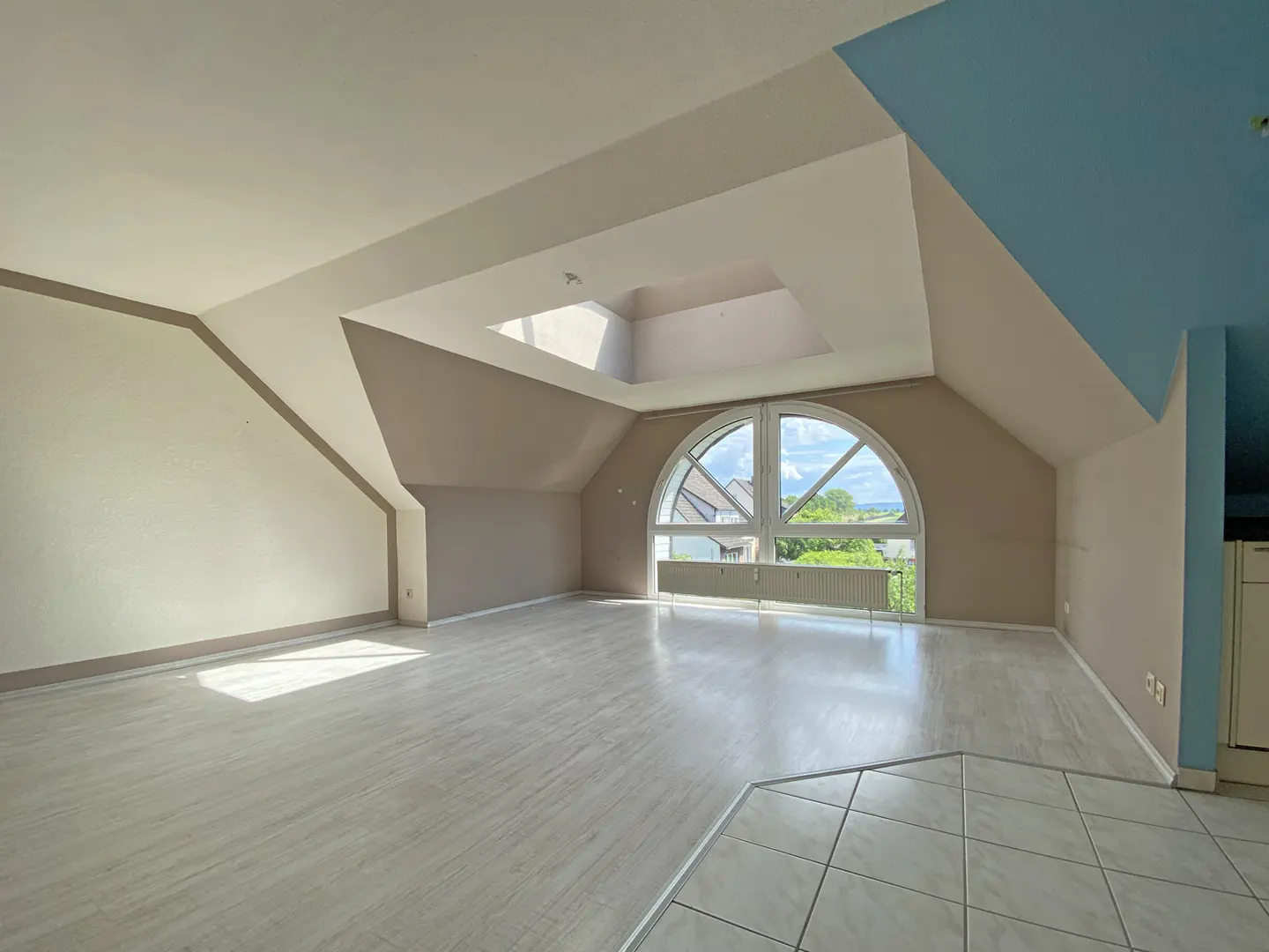 Empty attic room with light wood floors, beige walls, a blue accent wall, a skylight, and a large arched window.