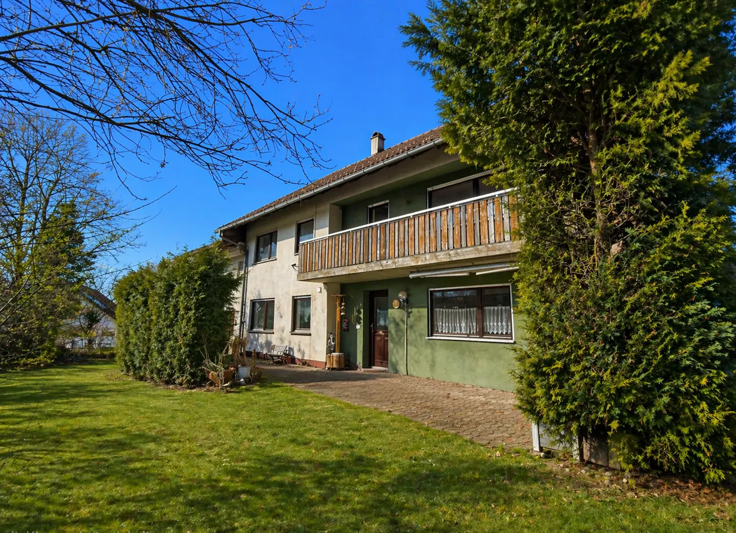Two-story house with a wooden balcony, green and beige exterior, surrounded by trees and a green lawn under a clear blue sky.