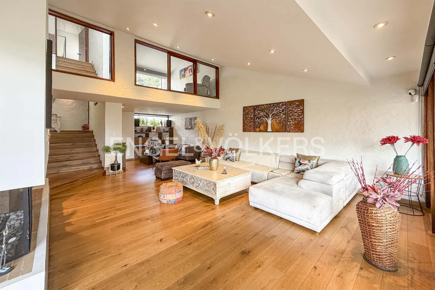 Bright living room with hardwood floors, a white sectional sofa, and a carved white coffee table. Stairs lead to a loft area with windows.
