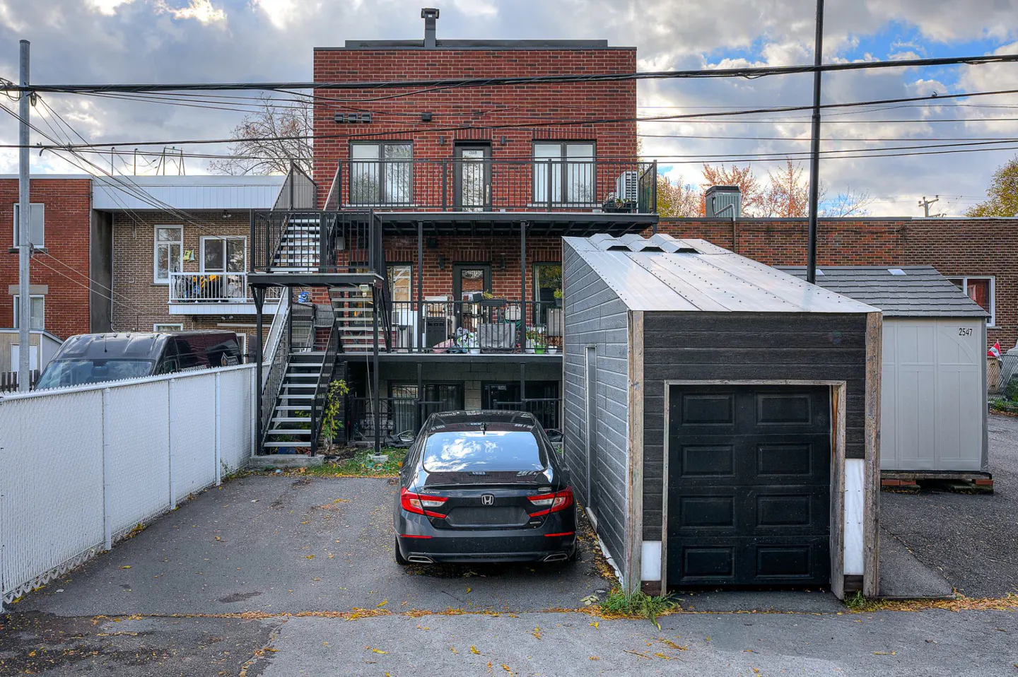 Backyard view of a three-story red brick apartment building with black metal stairs and balconies, a black car, and a black garage.