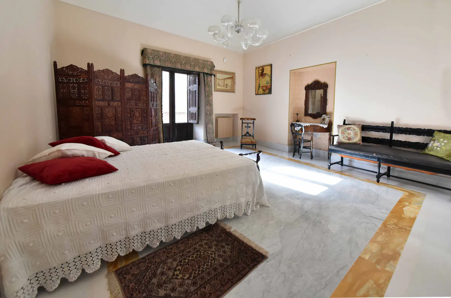 Bedroom with white bedspread, red pillows, and carved wooden screen. Marble floors, bench, and antique furniture. Chandelier.