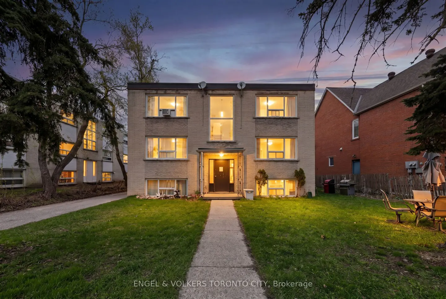 Two-story apartment building with a gray brick facade, illuminated windows, and a green lawn at dusk.