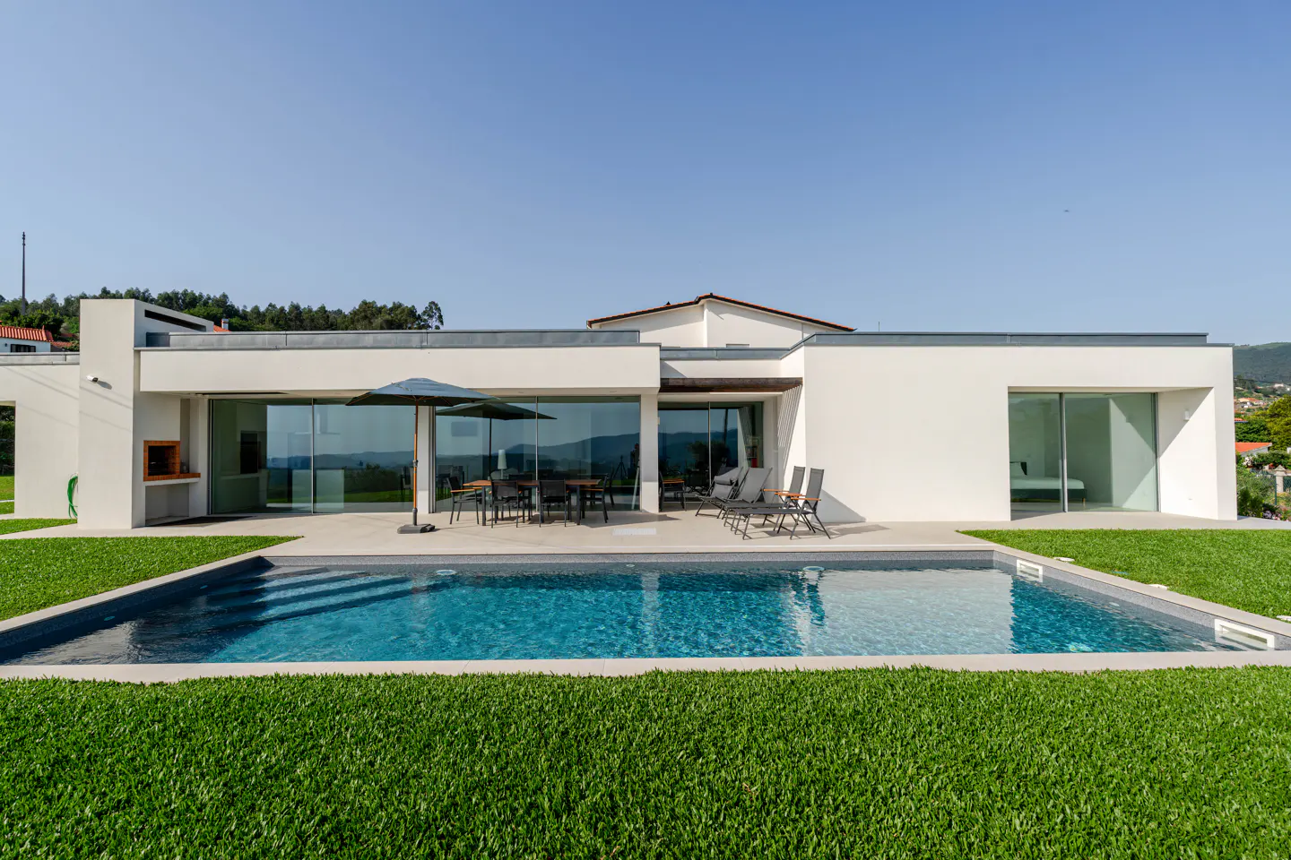 Modern white villa with a swimming pool, patio furniture, and large glass sliding doors under a clear blue sky.