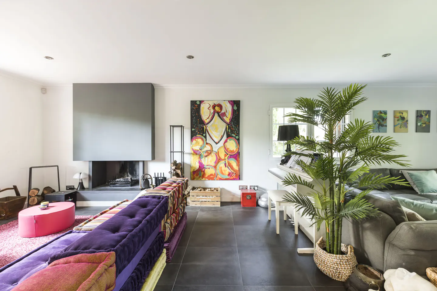 Bright living room with a gray fireplace, colorful art, and a purple couch. A white piano and potted palm add to the room's eclectic style.