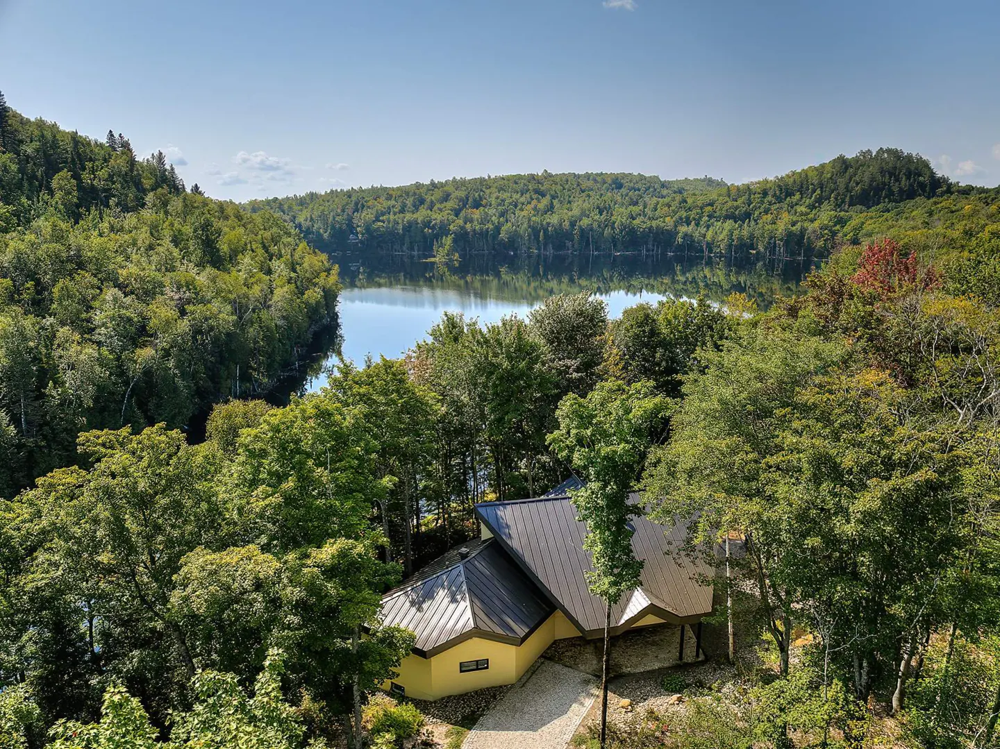 Aerial view of a yellow house with a black roof surrounded by green trees and a lake in the background.