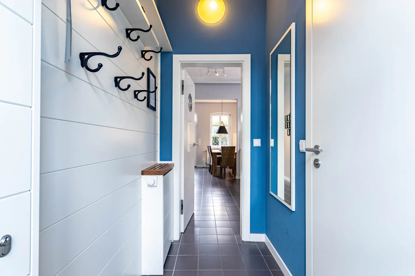 Hallway with blue walls, white trim, and gray tile floor. A white paneled wall has black coat hooks and a wooden bench. A mirror hangs on the blue wall.