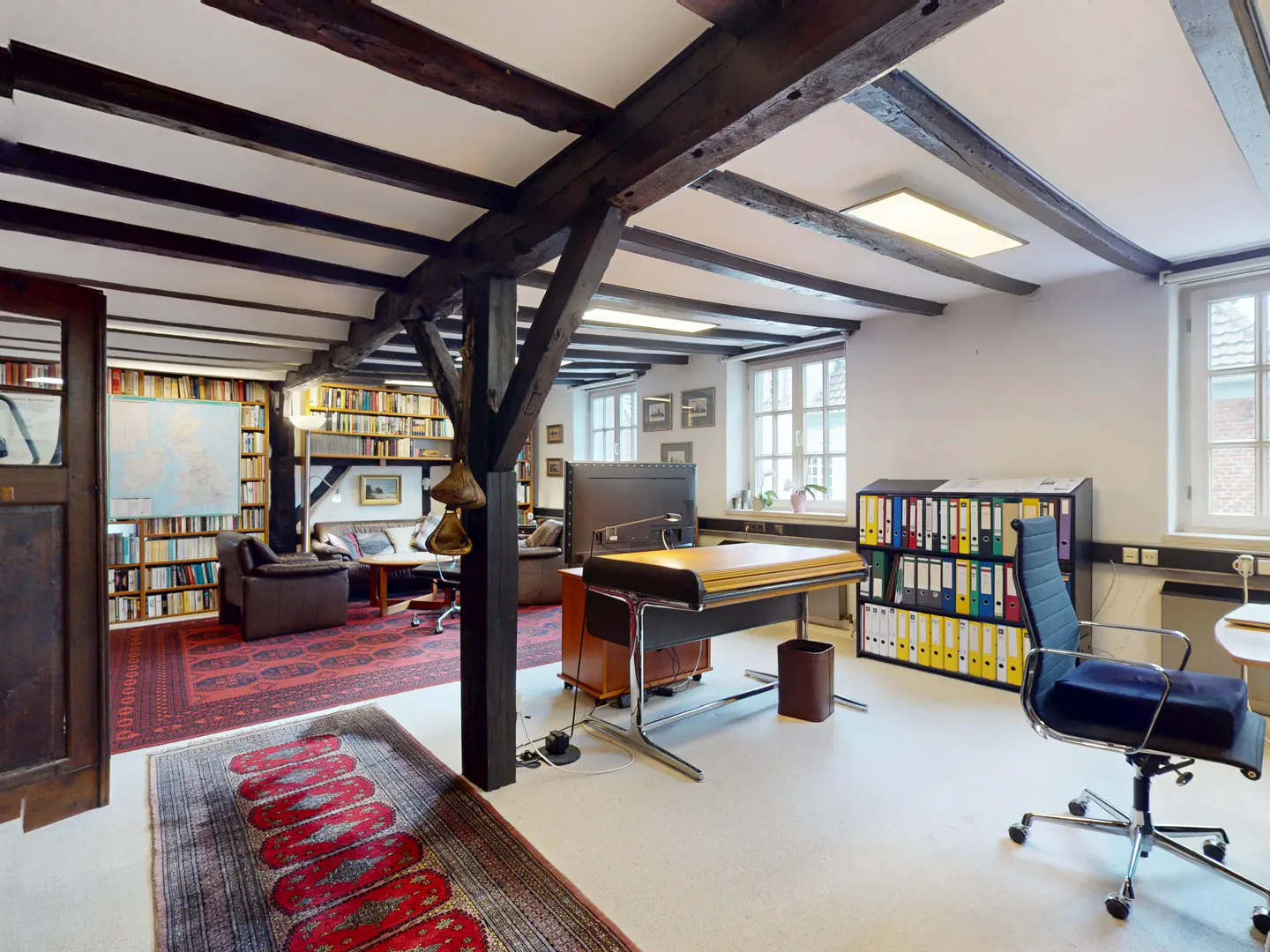 Office space with exposed beams, bookshelves, and a red rug. A desk and chair are in the foreground.