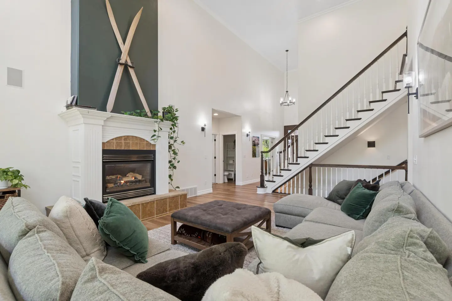 Living room with a gray sectional sofa, fireplace with crossed skis above, and a staircase with white railings.