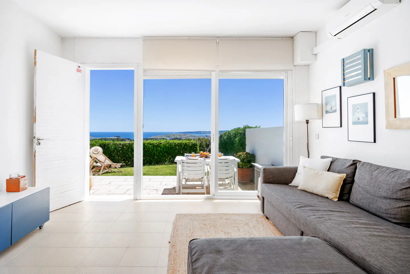 Bright living room with a gray sofa, white walls, and a view of the ocean through sliding glass doors. A table and chairs sit on the patio.