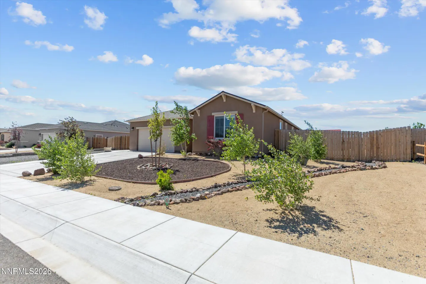 A single-story, tan house with red shutters, a two-car garage, and a brown mulch garden under a blue, cloudy sky.