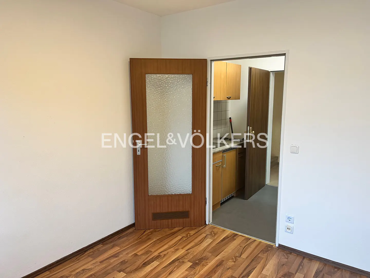 A room with wood-look flooring, white walls, and a wood door with textured glass. An open doorway leads to a small kitchen with wood cabinets.
