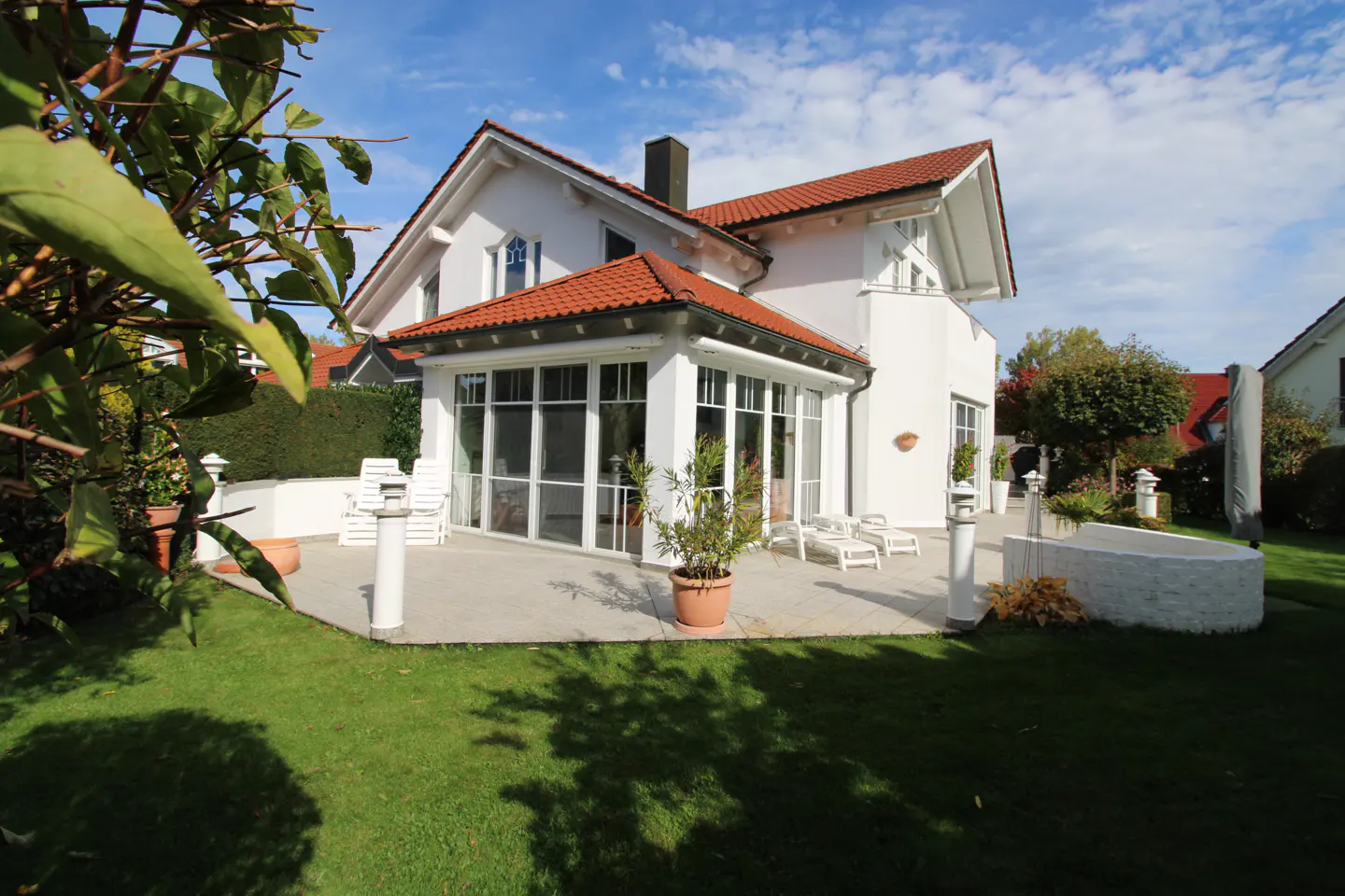 Backyard view of a white house with a red tile roof and a sunroom, surrounded by a green lawn and trees under a blue sky.