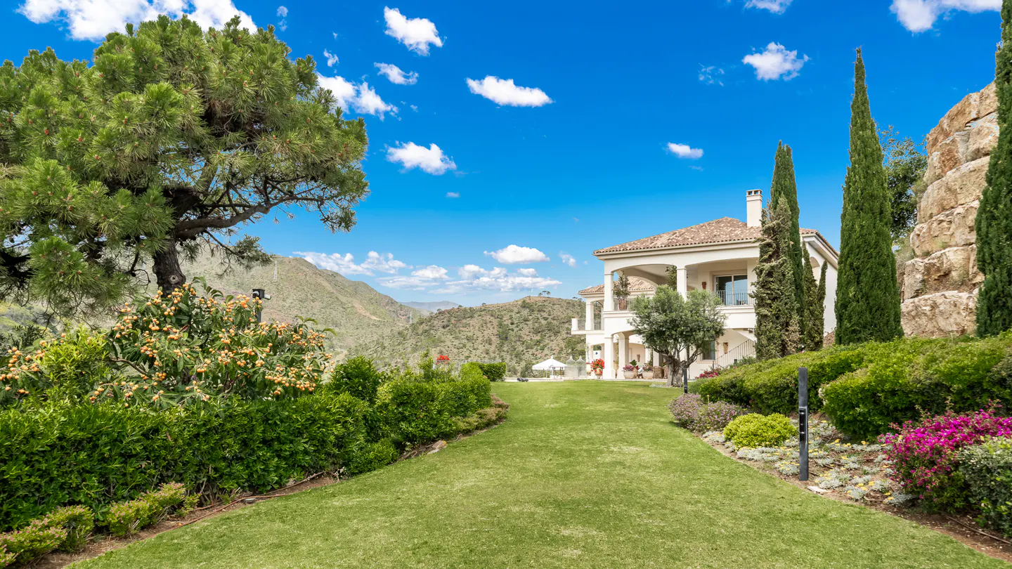 Luxury villa with a tile roof and white columns, surrounded by green lawns, trees, and a mountain view under a blue sky.