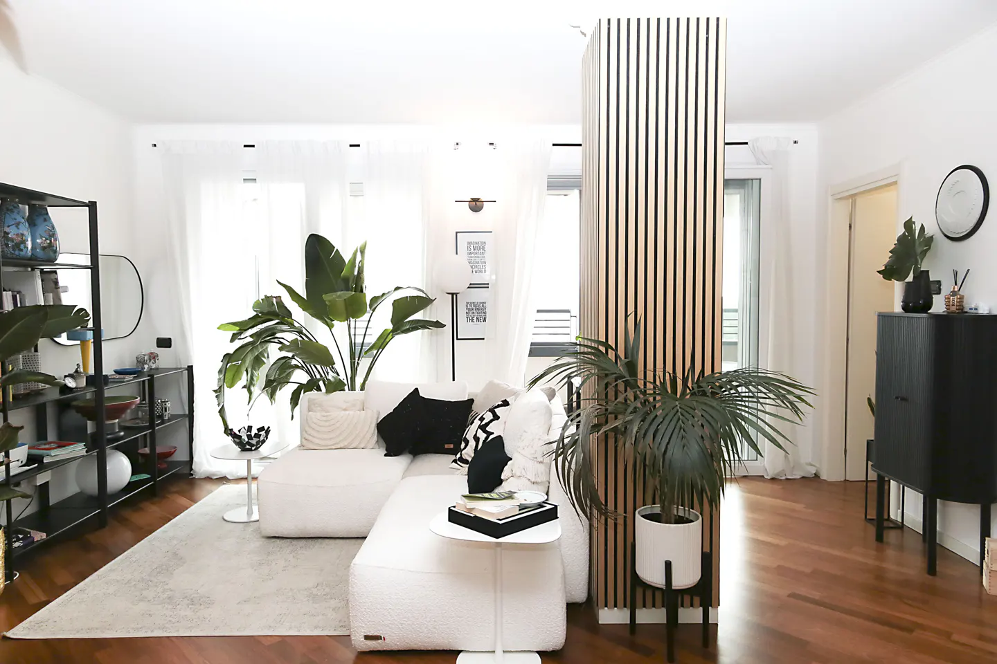 Bright living room with white sofa, wood floors, and plants. A wood slat wall divides the space. Black shelving and cabinet add contrast.