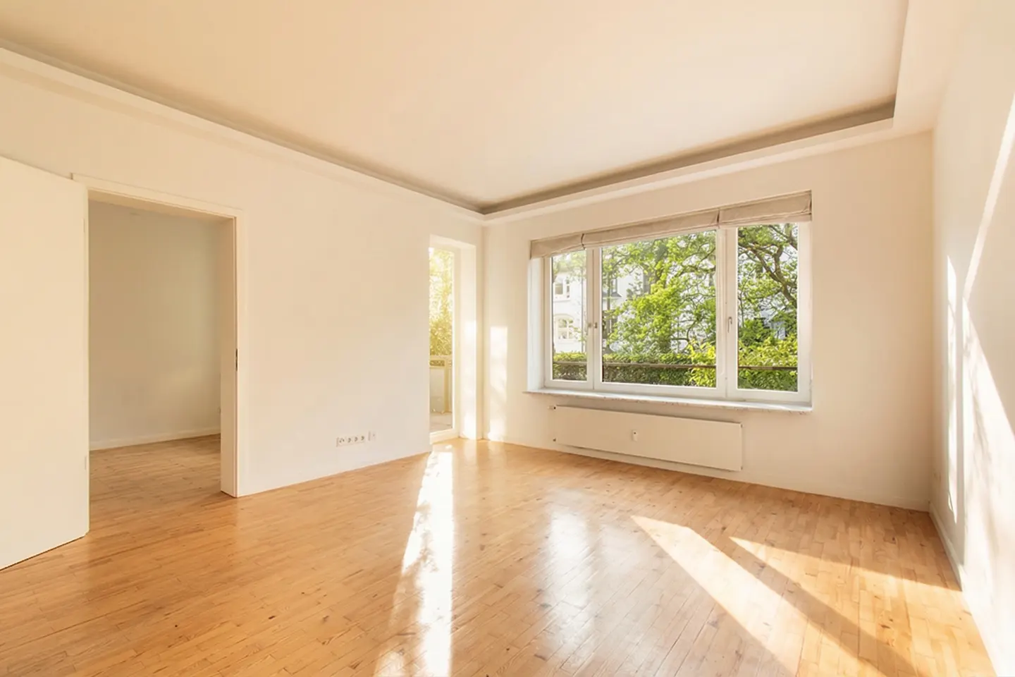 Bright, empty room with hardwood floors, white walls, and a large window overlooking green trees. A doorway leads to another room.