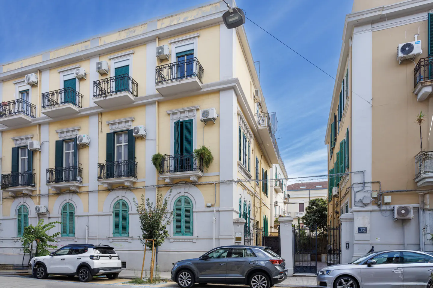 Street view of a yellow apartment building with green shutters and black wrought iron balconies. Cars are parked on the street.