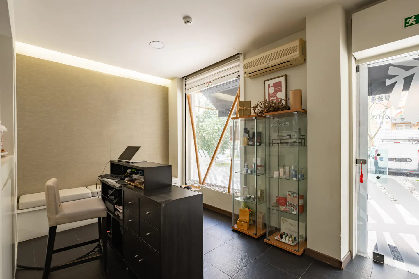 Bright, modern office space with a black desk, laptop, display cases, and a window view to the outside.