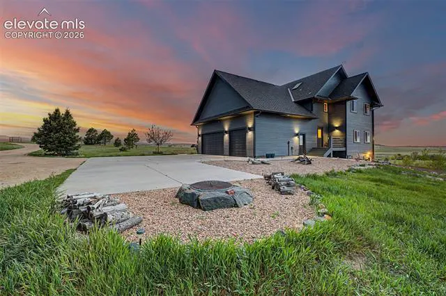 Modern dark gray two-story house with a concrete driveway, fire pit, and wood pile at sunset with a colorful sky.