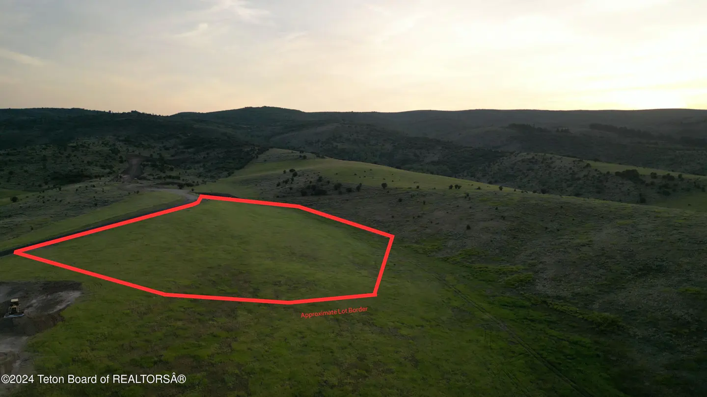 Aerial view of a green field outlined in red, indicating the approximate lot border, with rolling hills in the background.