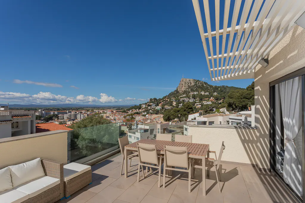 Balcony view with outdoor furniture and city skyline. Table, chairs, and sofa on a tiled patio with a mountain backdrop under a blue sky.