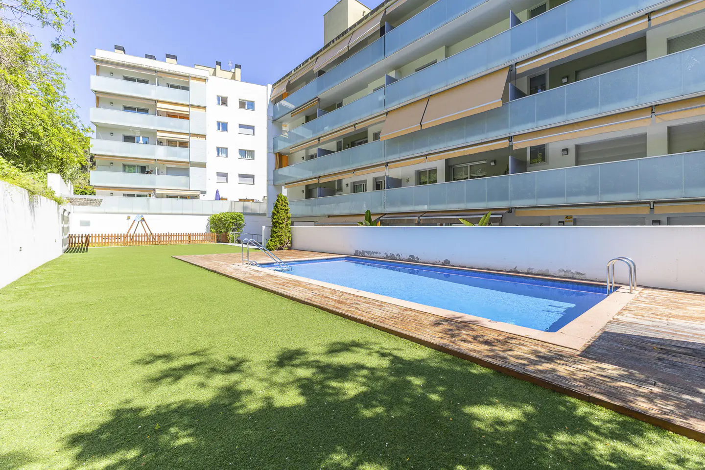Apartment complex with a blue swimming pool, wooden deck, and green lawn. Balconies with glass railings are visible.