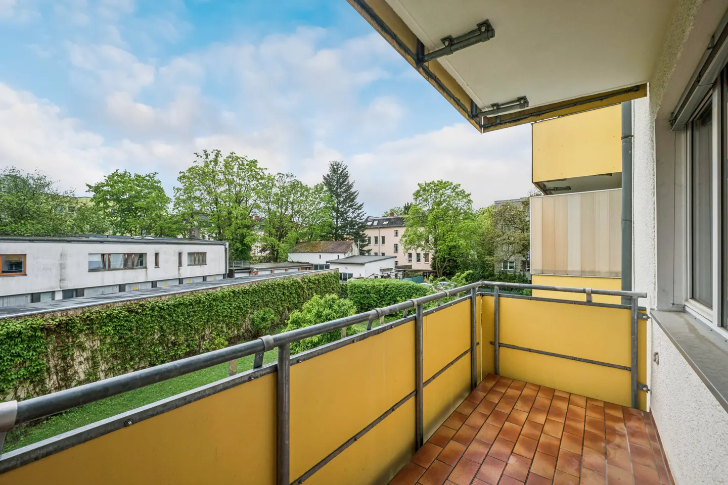 Balcony view with yellow panels and metal railing overlooking green trees and buildings under a cloudy sky. Brown tiled floor.