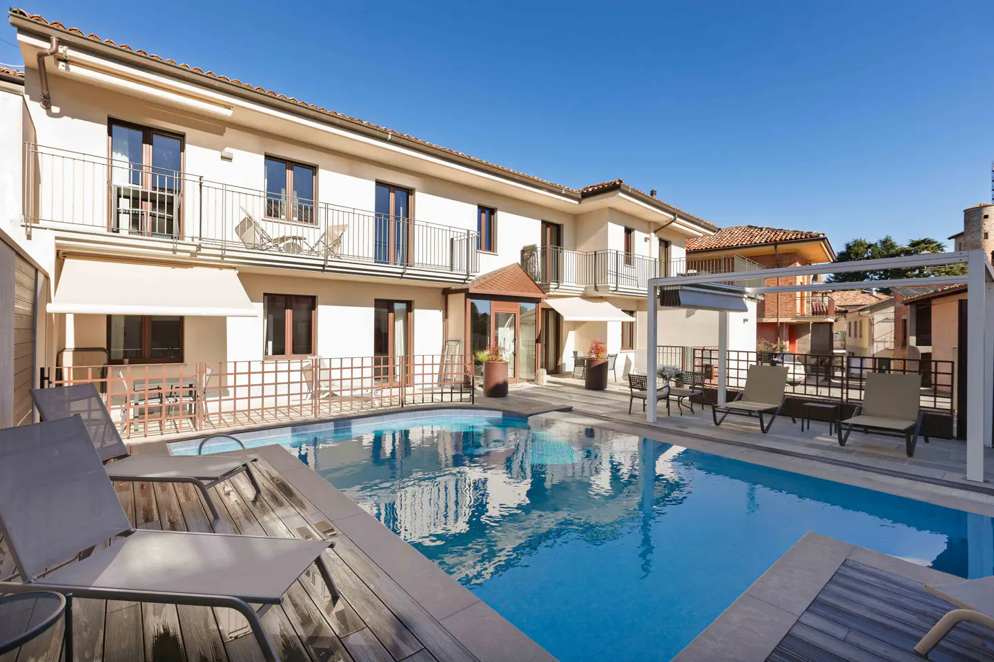 A modern two-story villa with a swimming pool, wooden deck, lounge chairs, and balconies under a clear blue sky.