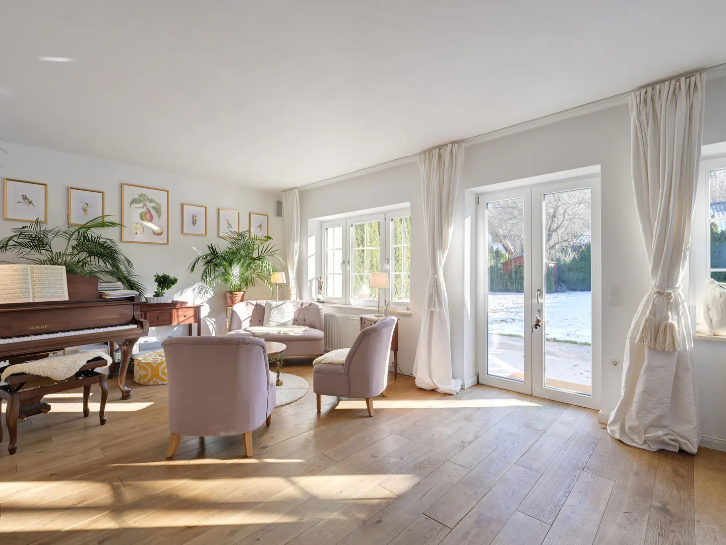 Bright living room with hardwood floors, piano, and lilac armchairs. White walls feature framed art and windows with white curtains. Double doors lead to a snowy yard.