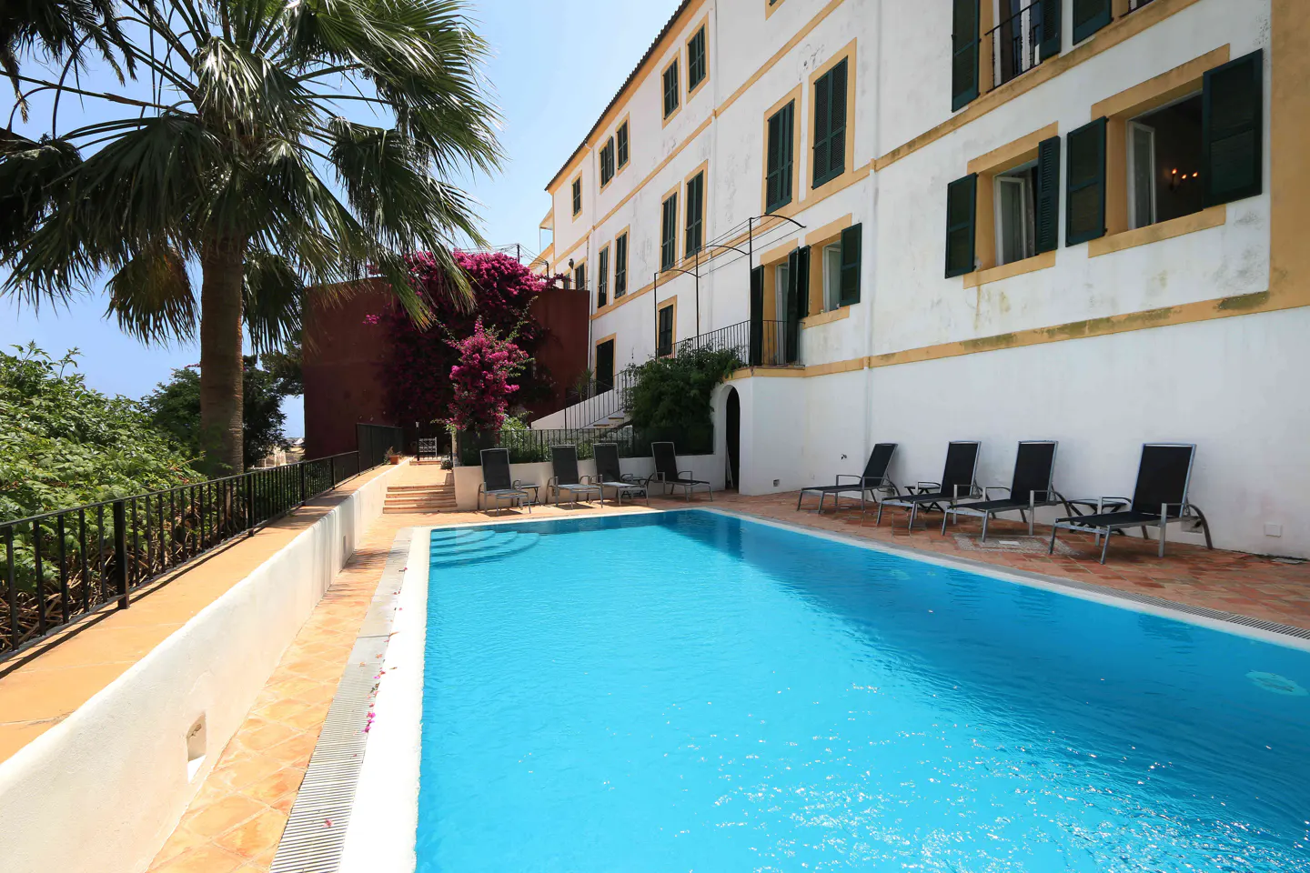Outdoor pool with lounge chairs next to a white building with green shutters and a palm tree.