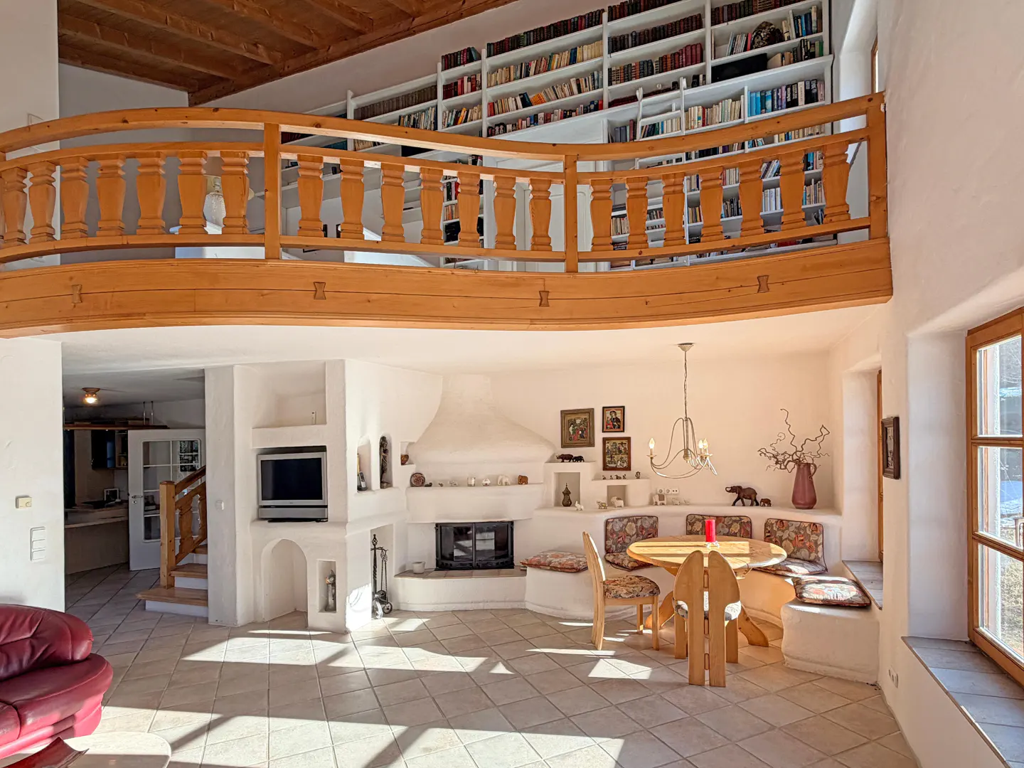 Bright living room with a wooden balcony overlooking a fireplace, dining area, and a floor-to-ceiling bookshelf.