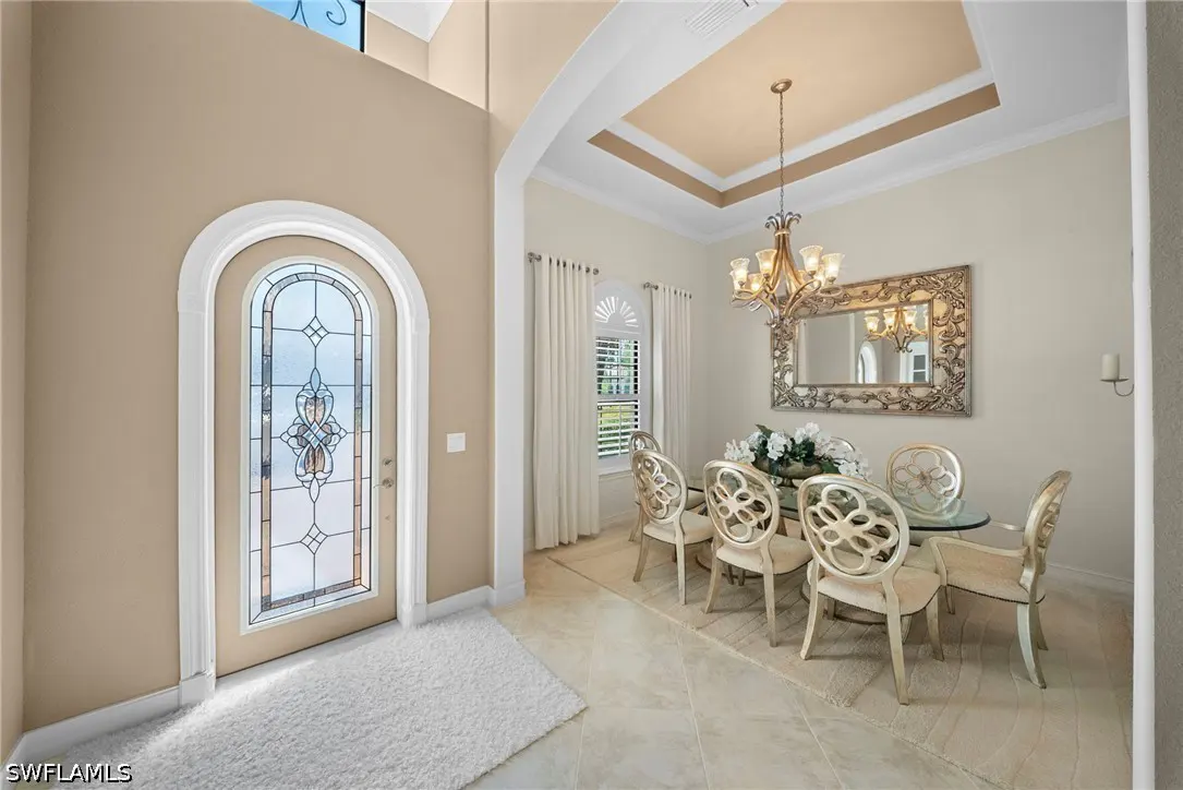 A bright foyer and dining room with a glass table, ornate chairs, and a decorative chandelier. A patterned glass front door is on the left.